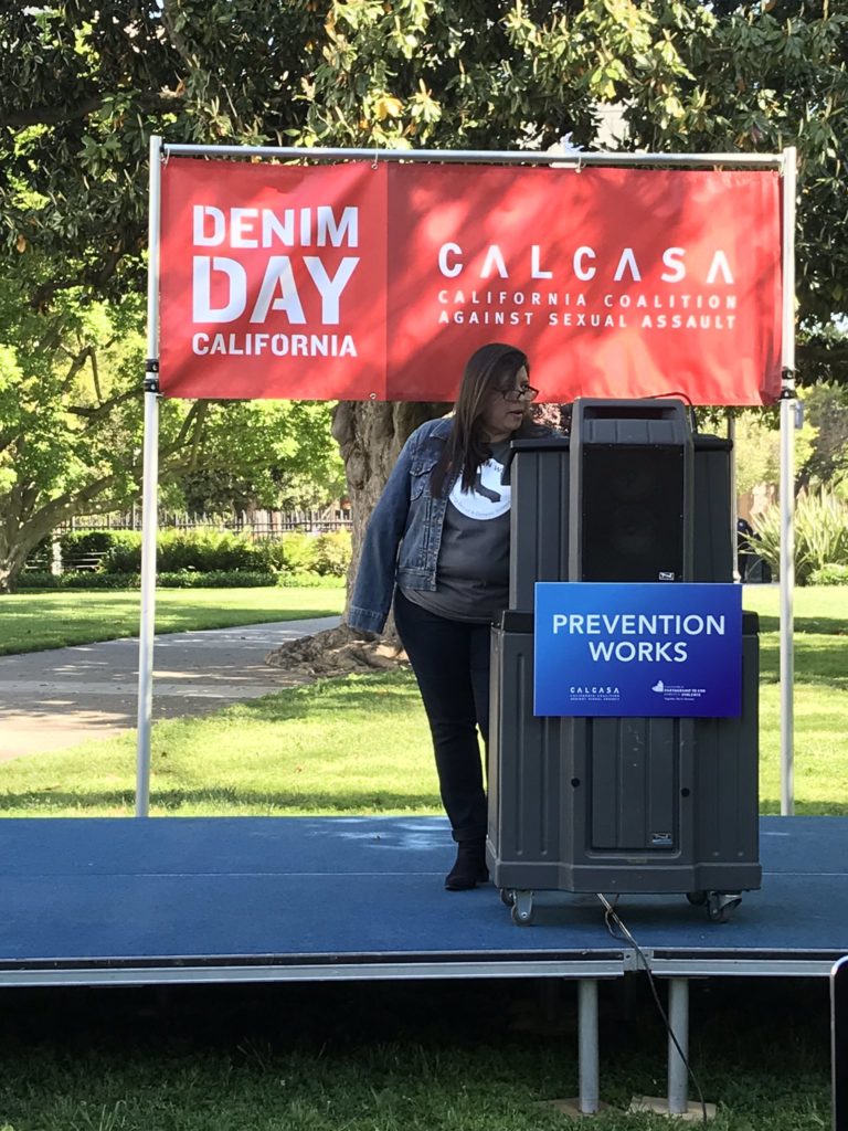 Red banner with white writing daying Denim Day and with CALCASA Logo in background. Latina women wearing denim jacket at podium with sign saying Prevention Works